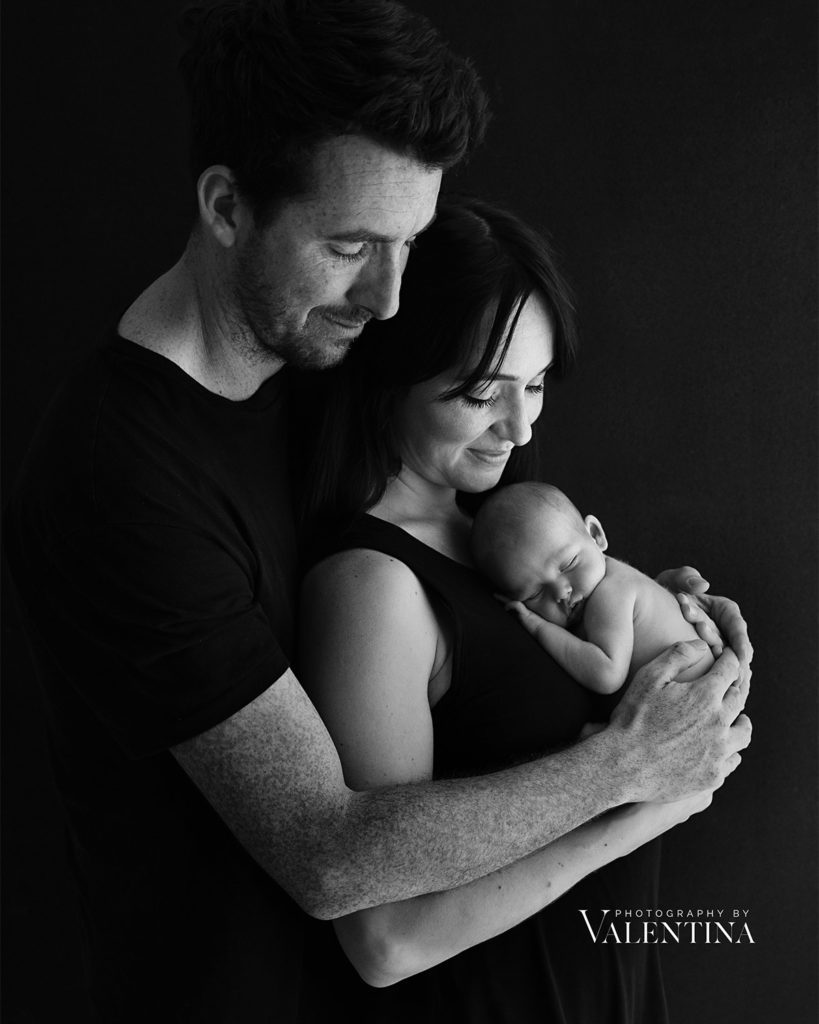 Why Hiring a Professional Newborn Photographer Matters: black and white portrait of new parents lovingly holding their newborn baby in a calm and intimate studio setting by Photography by Valentina.
