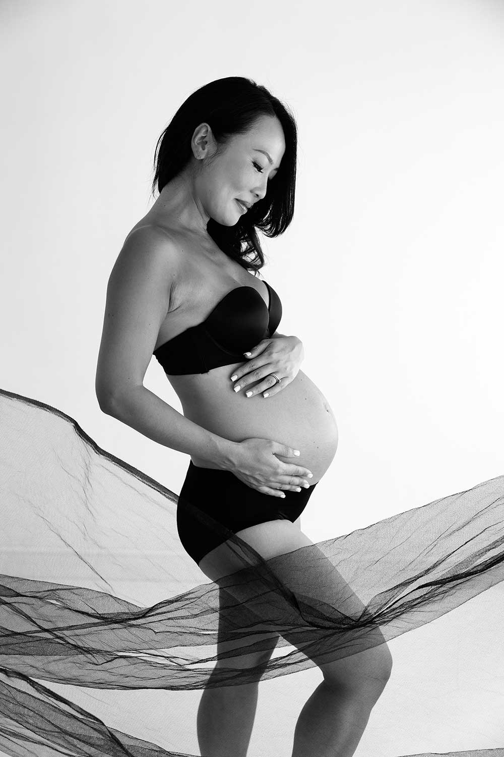 Black and white studio maternity portrait of a woman wrapped in sheer fabric.