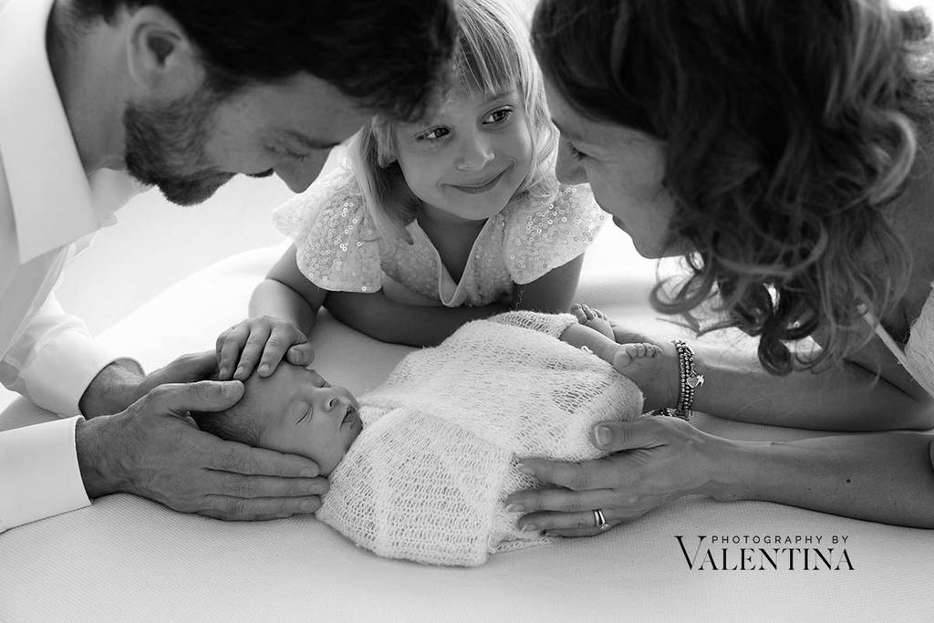 black and white family photo. Newborn baby is laying on a bean bag and parents and sister are holding their hands around him 